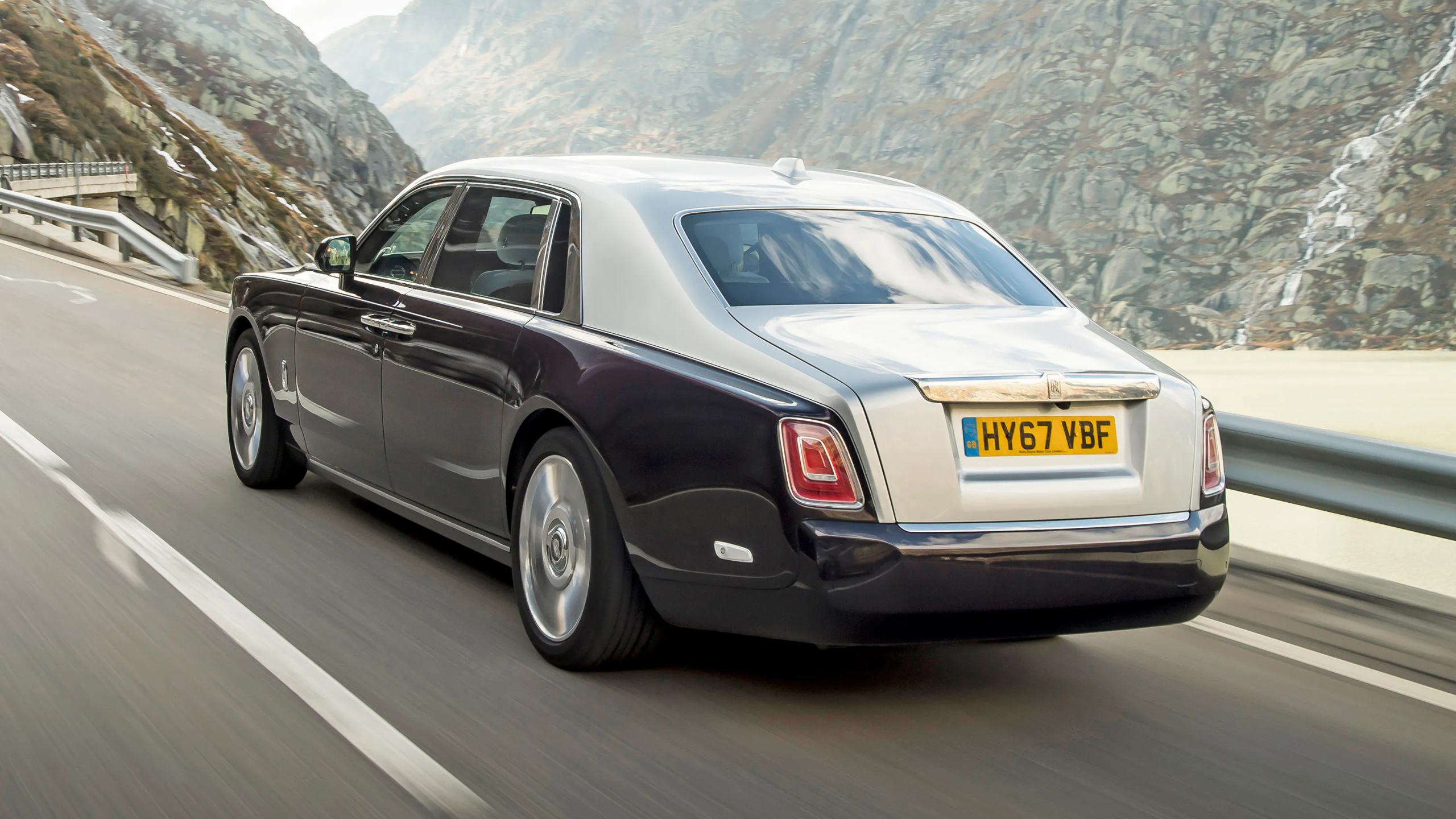 Three-quarter rear view of a 2026 Rolls-Royce Phantom cruising through a scenic mountain highway
