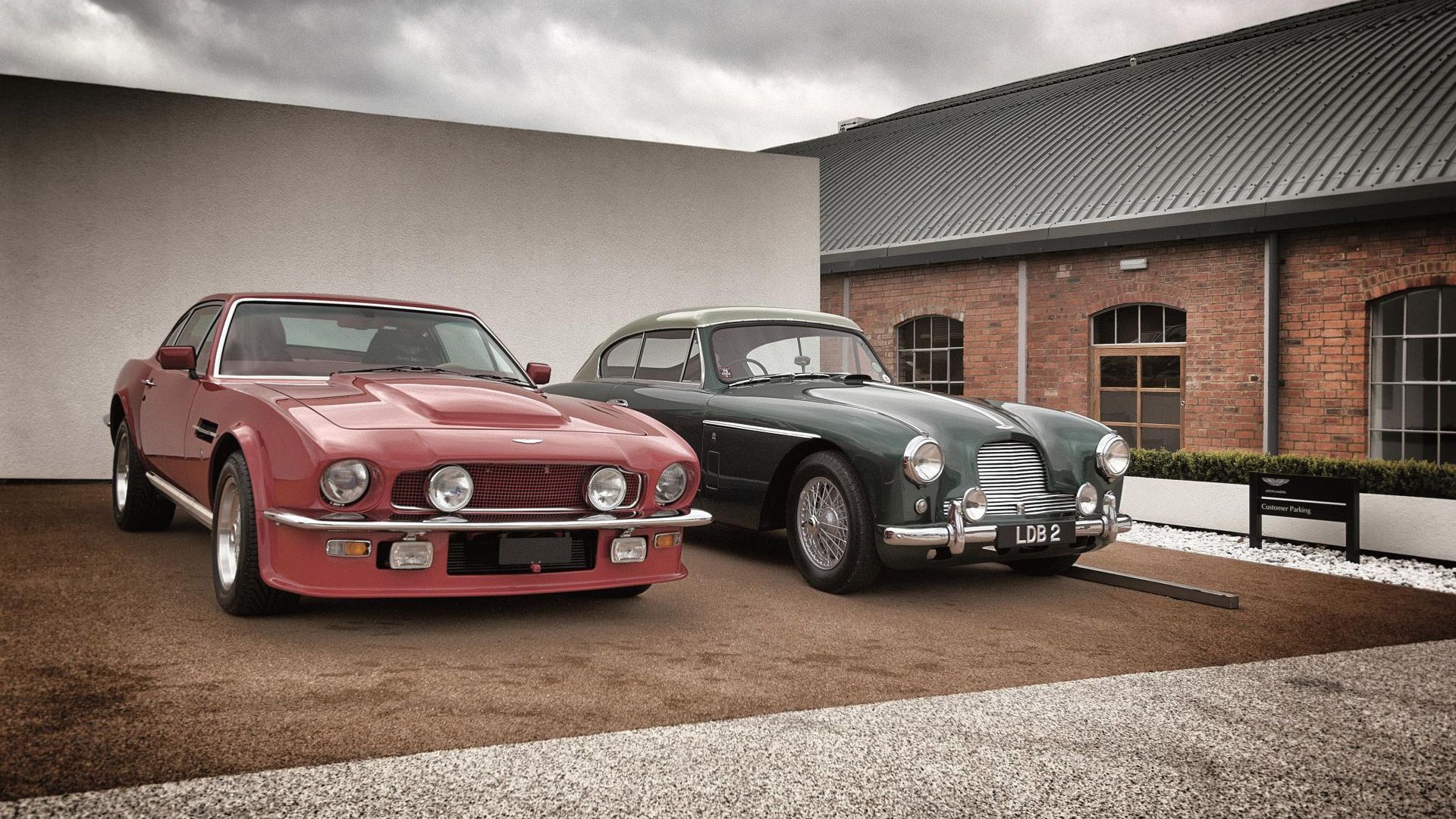 Two vintage Aston Martins, a red V8 Vantage and a green DB2, parked side-by-side in front of a brick building.