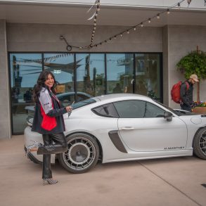 Salena from Porsche Monterey waves the checkered flag while showing off her race attire in front of a Porsche GT4 RS.