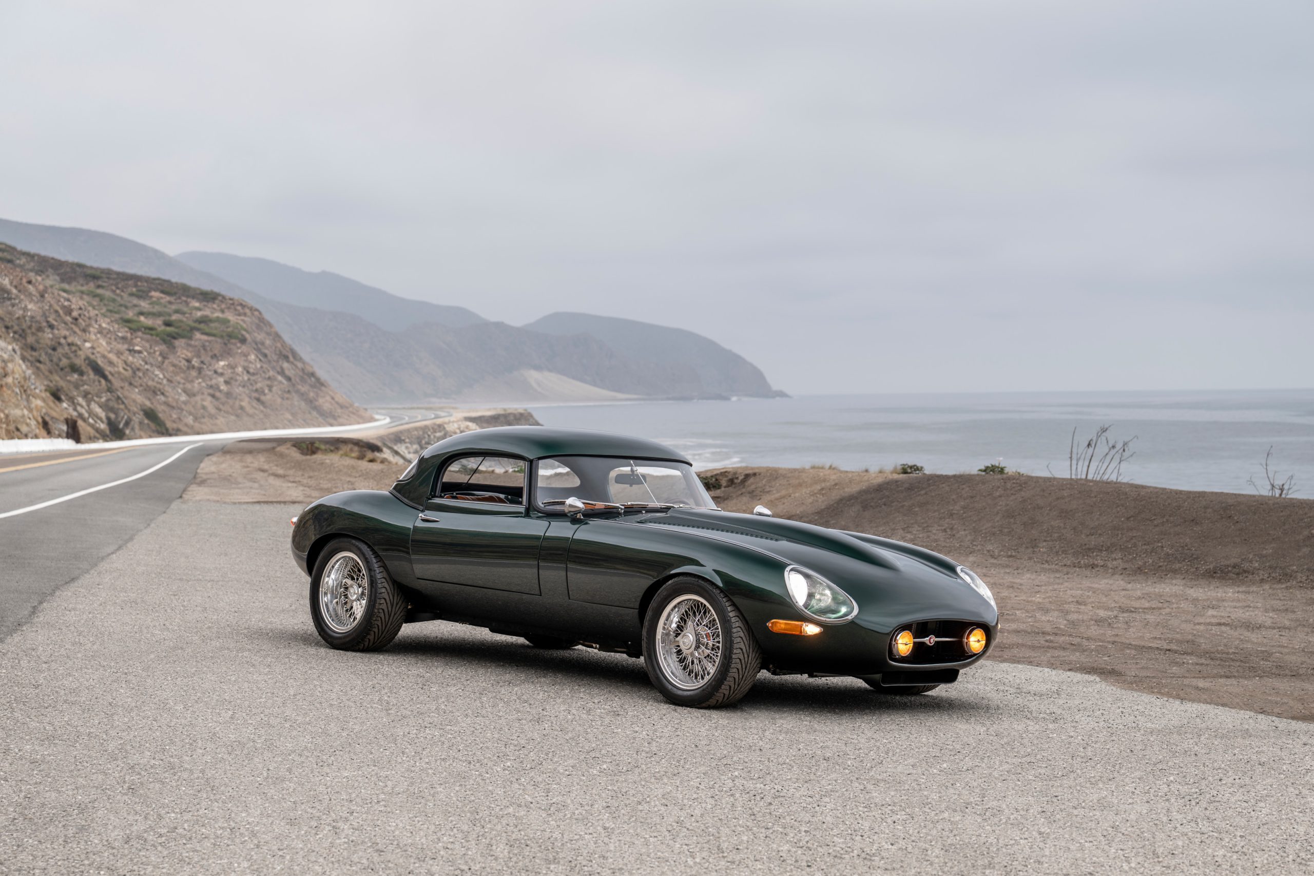 Three-quarter front view of a green Jaguar E-Type with coastline background